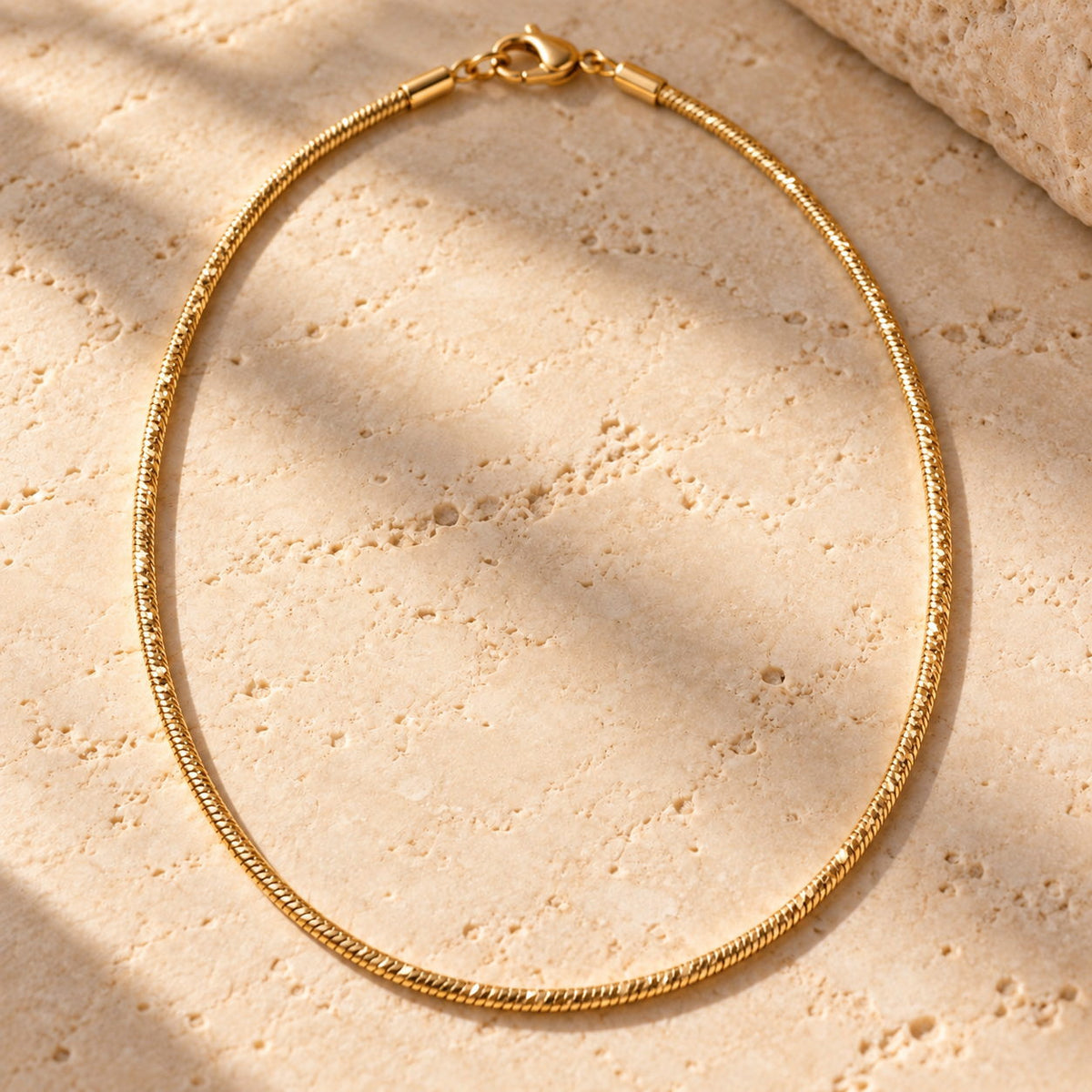 Bar None diamond-cut gold snake chain necklace on travertine, facets catching the light